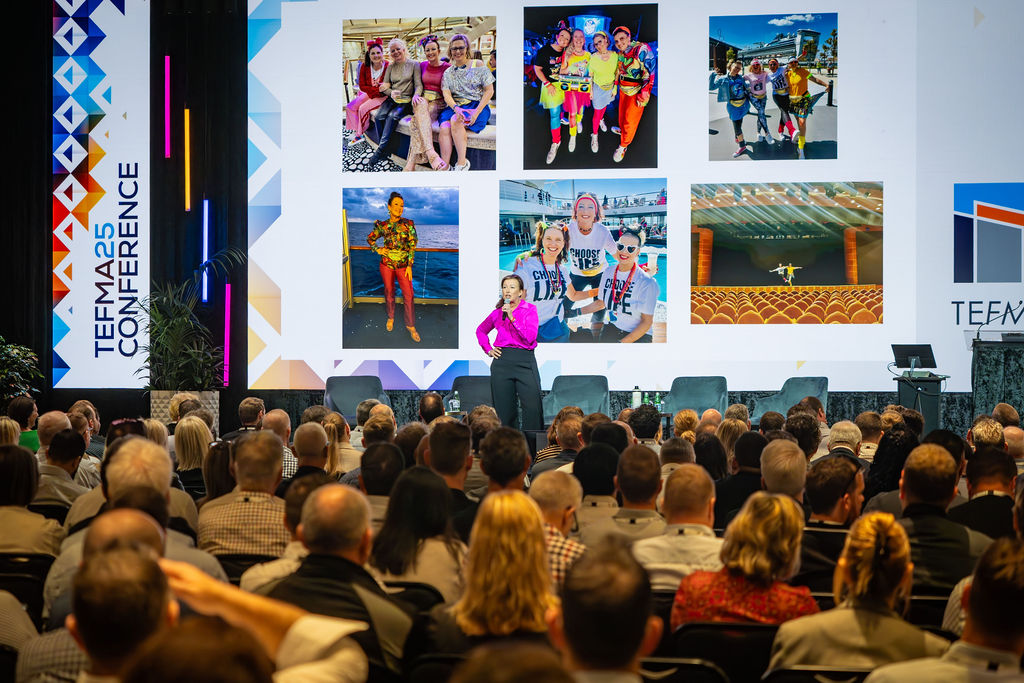 A speaker presenting to a packed conference audience beneath a large photo collage screen